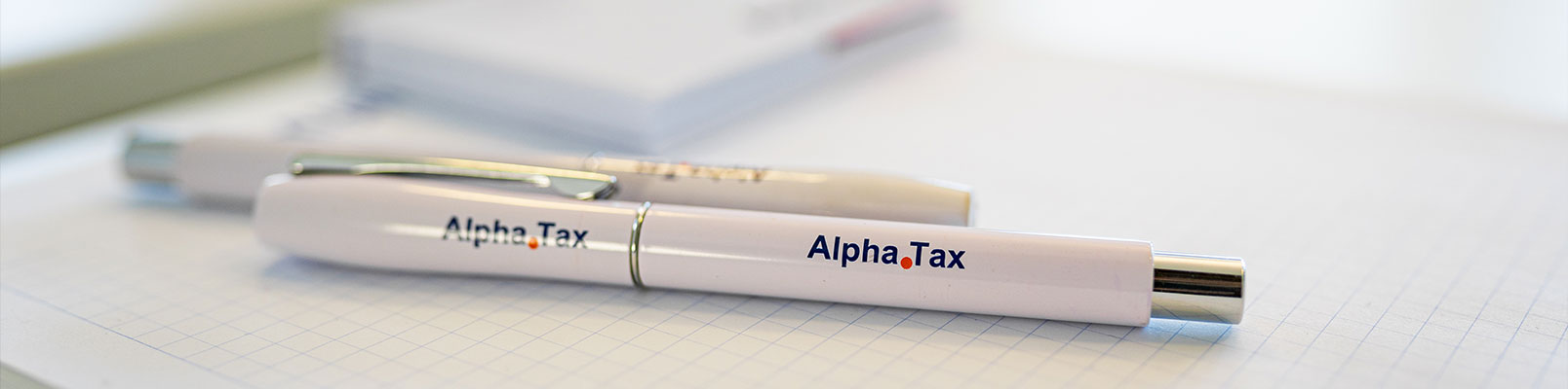 Kugelschreiber mit AlphaTax-Logo auf einem karierten Notizblock in einer Büroumgebung.