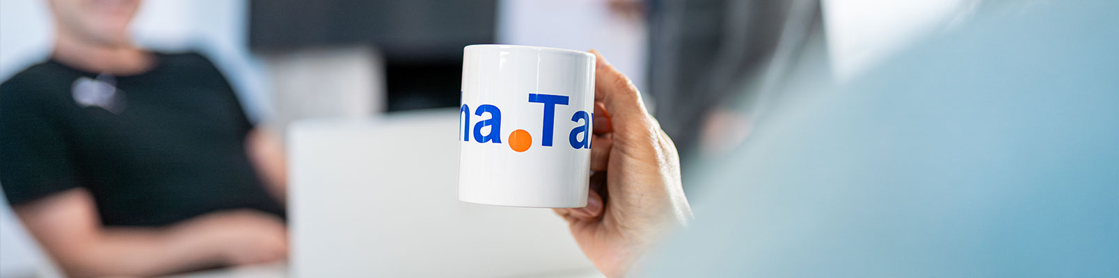 Hand hält eine Tasse mit AlphaTax-Logo während eines Gesprächs im Büro.